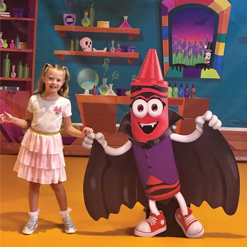 Child standing next to a red crayon character in a vampire costume